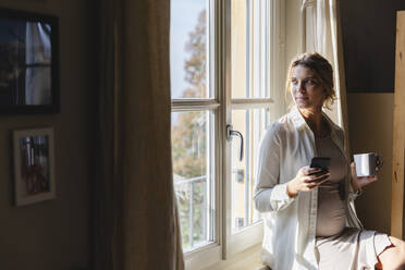 Junge schwangere Frau mit Kaffeetasse und Handy in der Hand, während sie durch das Fenster ihres Hauses schaut - MRAF00606