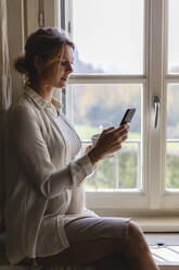 Pregnant woman using mobile phone while sitting by window - MRAF00608