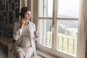 Schwangere Frau, die mit ihrem Handy telefoniert und durch ein Fenster schaut - MRAF00610