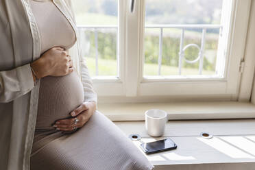 Pregnant woman hands on stomach sitting by window at home - MRAF00612