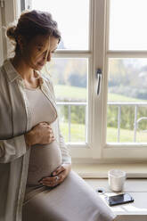 Pregnant woman holding her belly by window - MRAF00613