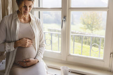 Young pregnant woman holding her belly by window at home - MRAF00614