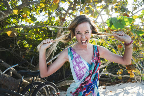 Cheerful woman screaming while pulling hair against bush - JMPF00713