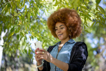 Smiling woman with frizzy hair using mobile phone at public park - OCMF01865