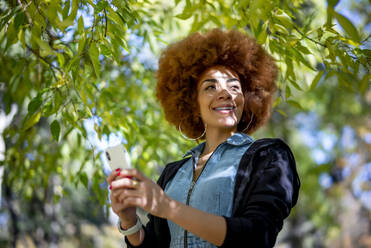 Smiling woman wearing hoop eating while holding mobile phone at public park - OCMF01866