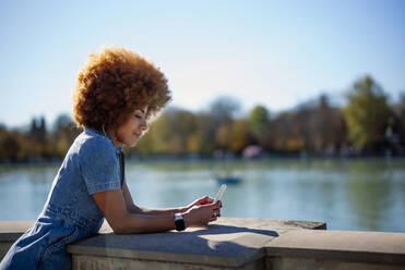 Mid adult woman with frizzy hair using mobile phone at Estanque Grande Del Retiro pond on sunny day, Spain - OCMF01869