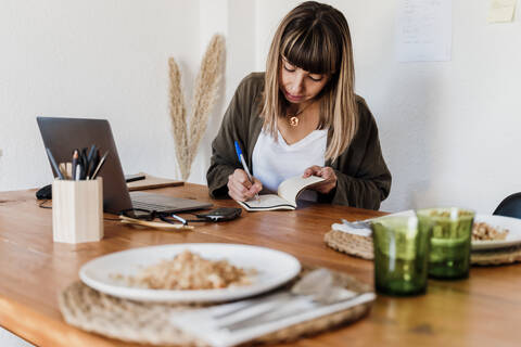 Female professional with food plate on table writing on notepad in home office stock photo