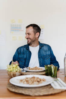 Glücklicher männlicher Berufstätiger mit Essen auf dem Tisch, während er im Wohnzimmer wegschaut - AFVF07837