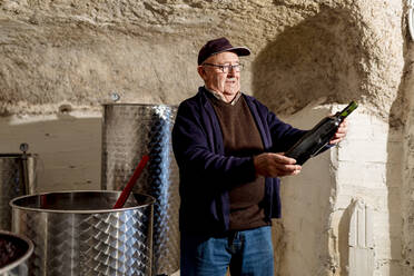 Älterer männlicher Winzer mit einer Flasche Rotwein in einem Weingut - EGAF01268