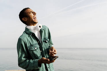 Cheerful man laughing while holding mobile phone against sea and sky - AFVF07865