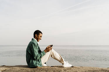 Happy man text messaging through smart phone while sitting on pier by sky and sea - AFVF07868