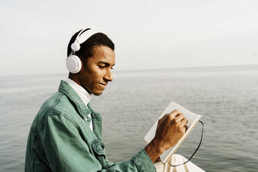 Smiling man writing in book by sea and sky - AFVF07870