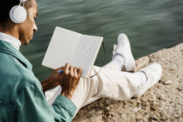 Junger Mann zeichnet in einem Buch, während er auf einem Pier am Meer sitzt - AFVF07871
