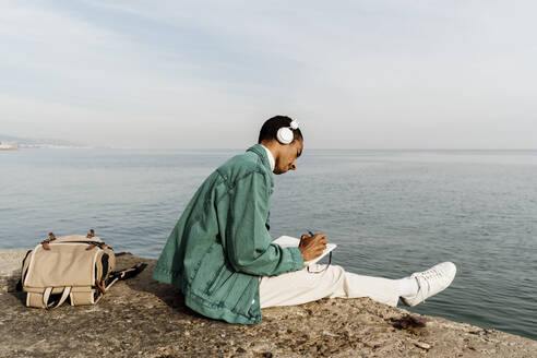 Junger Mann mit Kopfhörern schreibt in Tagebuch am Meer und Himmel - AFVF07873