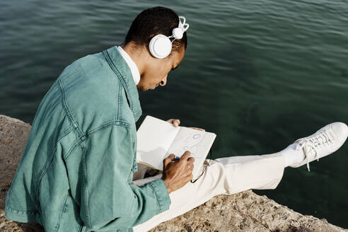 Mann zeichnet in einem Buch, während er auf einem Pier am Meer sitzt - AFVF07874