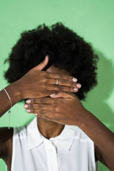 Young woman hiding face with hand while standing against green background - GIOF09948