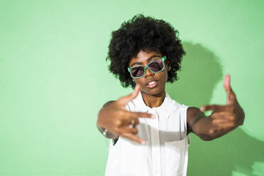 Woman wearing sunglasses gesturing while standing against green background - GIOF09954