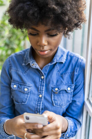 Young woman using mobile phone while leaning on window at home stock photo