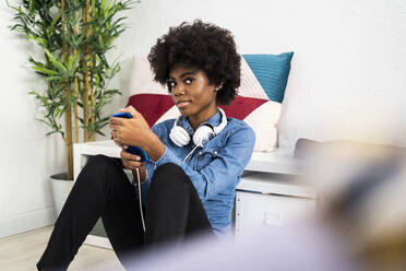 Woman with mobile phone and headphones staring while sitting at home - GIOF10005