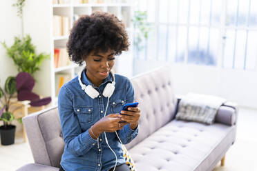 Frau mit Kopfhörern, die ein Mobiltelefon benutzt, während sie zu Hause auf dem Sofa sitzt - GIOF10007