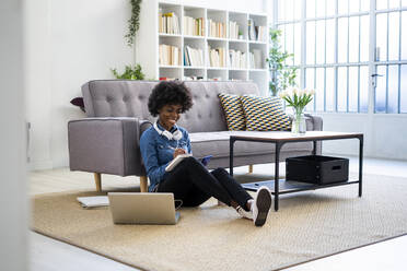 Smiling woman writing in book while sitting by laptop on floor at home - GIOF10011