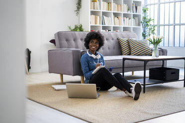 Thoughtful woman with book looking away while sitting by laptop on floor at home - GIOF10012