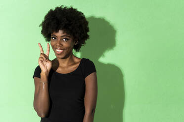 Smiling woman showing peace gesture while standing against green background - GIOF10026