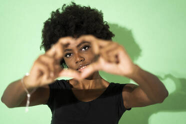 Woman making heart shape with finger while standing against green background - GIOF10030