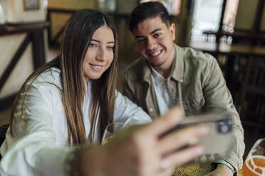 Teenage girl taking selfie on smart phone while sitting by male friend in bar - EGAF01296