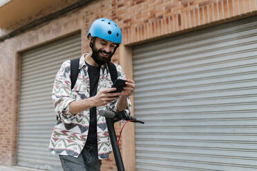 Young man with electric push scooter using smart phone while standing by shutter - EGAF01314