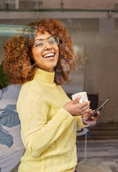 Lachende Frau mit Mobiltelefon beim Kaffee trinken hinter Glas in einem Cafe - VEGF03342