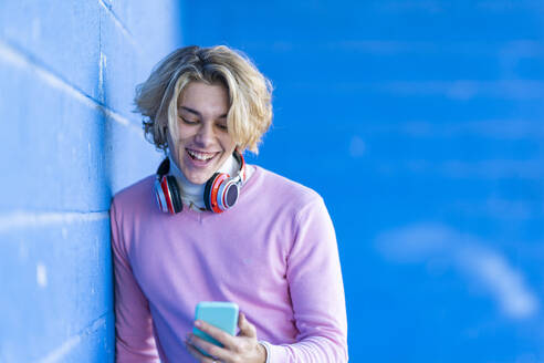 Happy fashionable young man using mobile phone while leaning on blue wall - GGGF00481