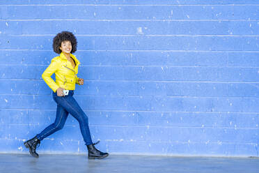 Glückliche Frau in gelber Lederjacke, die ihr Handy hält, während sie gegen eine blaue Wand läuft - GGGF00492