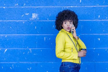 Afro young woman with surprised reaction talking on phone call against blue wall - GGGF00496