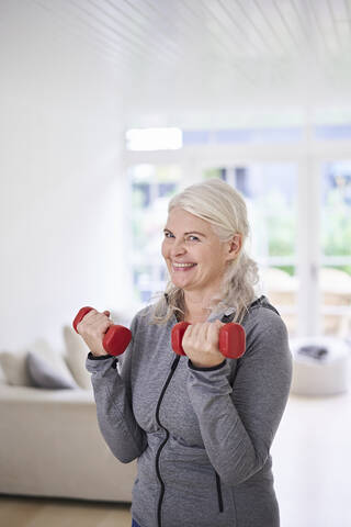 Smiling senior woman lifting dumbbells while exercising at home stock photo