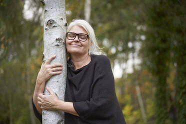 Lächelnde ältere Frau mit geschlossenen Augen, die einen Baumstamm in einem öffentlichen Park umarmt - JAHF00012