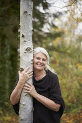 Happy senior woman hugging tree trunk in autumn park - JAHF00013