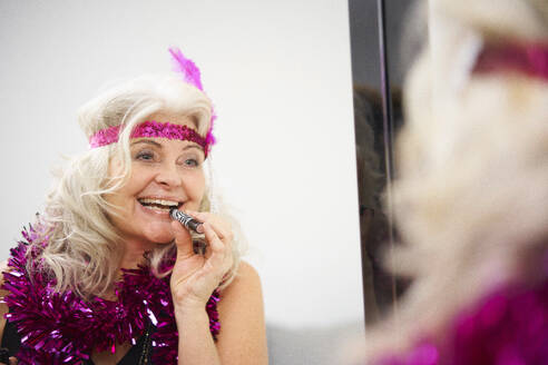 Happy senior woman with garland around neck applying lipstick in front of mirror at home - JAHF00020