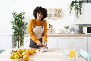 Smiling woman squeezing orange in bowl while standing at kitchen - GIOF10042