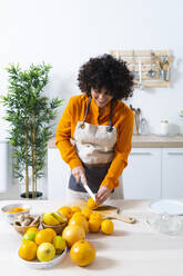 Young woman cutting orange for making juice while standing in kitchen at home - GIOF10043
