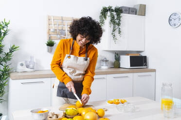 Woman smiling while cutting orange standing in kitchen at home - GIOF10045