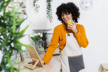 Smiling woman wearing apron drinking orange juice while using digital tablet at home - GIOF10053