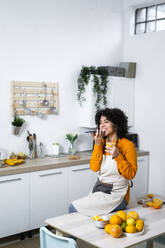 Lächelnde Frau mit Orangensaft, die mit ihrem Handy telefoniert, während sie zu Hause auf einem Tisch mit Obst sitzt - GIOF10059