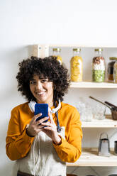 Young woman smiling while using mobile phone standing at home - GIOF10070