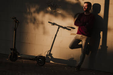 Mature man standing with arms crossed against wall during sunset - GMLF00927