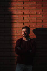 Man with arms crossed standing against brick wall during sunset - GMLF00928