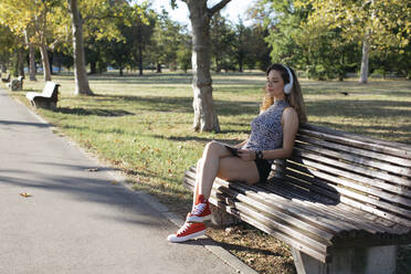 Young woman with eyes closed listening music through headphones in public park on sunny day - MOMF00920