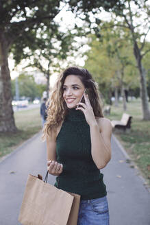 Lächelnde Frau, die wegschaut, während sie in einem öffentlichen Park telefoniert - MOMF00922