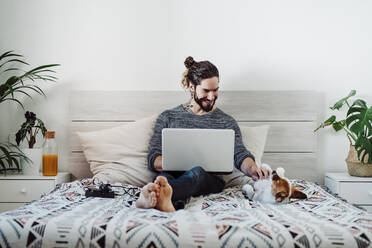 Lächelnder Mann, der mit seinem Hund spielt, während er einen Laptop zu Hause benutzt - EBBF01772