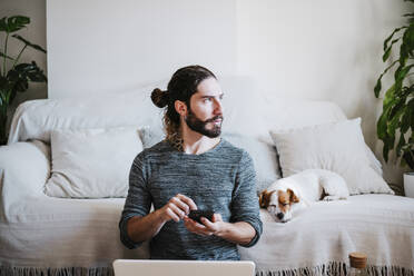 Mann mit Laptop und Mobiltelefon, der wegschaut, während er neben einem schlafenden Hund auf dem Sofa zu Hause sitzt - EBBF01775